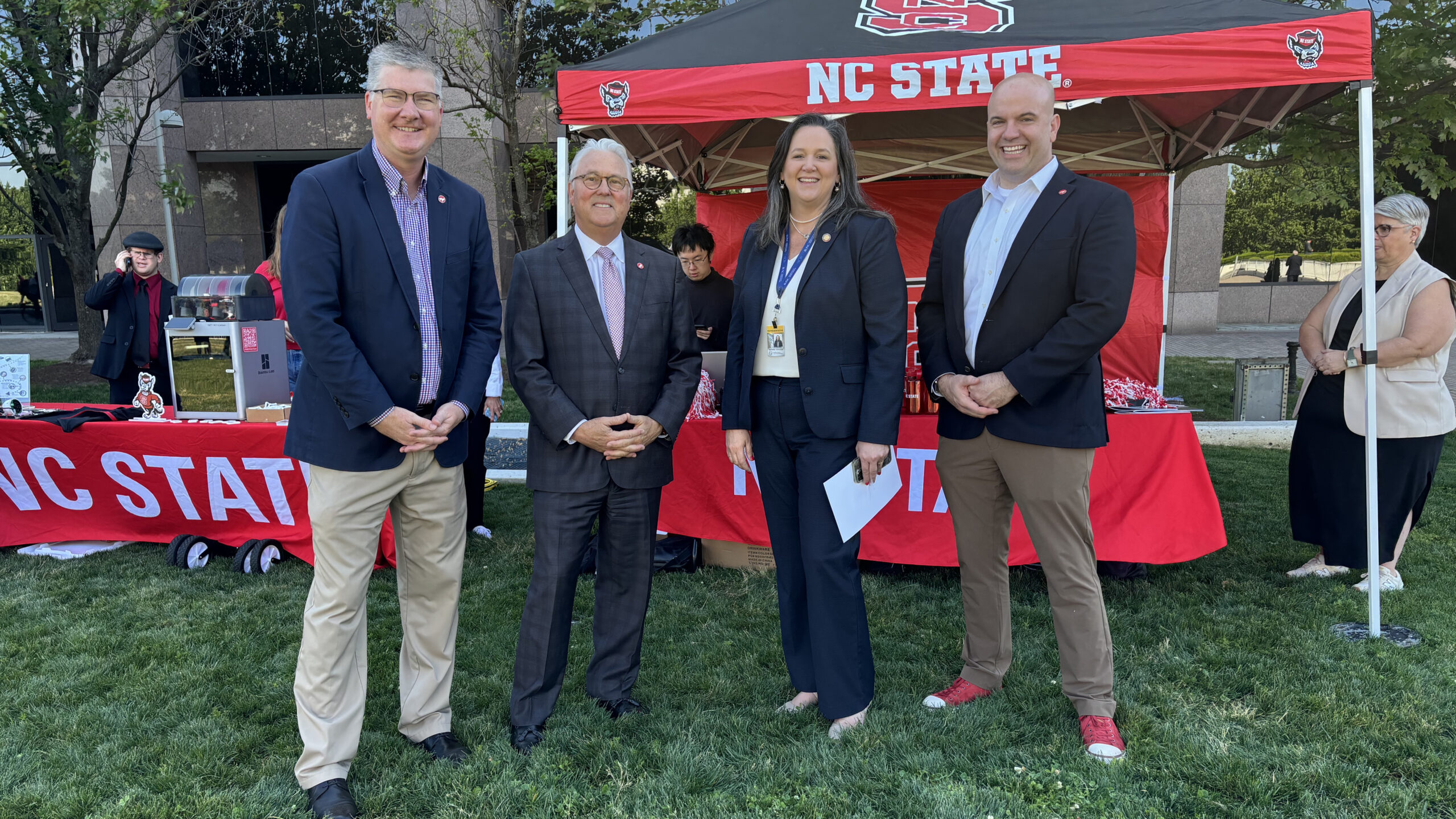 CALS dean Garey Fox, outgoing NC State chancellor Randy Woodson, an unidentified woman, and COE dean Jim Pfaendtner pose for a photo in front of a NC State table and canopy decorated in red, black and NC State logo.