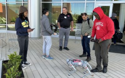 Five individuals are gathered around a small, light-colored robot dog on a paved outdoor area, with a modern building featuring large windows in the background. From left to right: a person in a dark beanie and dark sweatshirt is looking at a phone; a person in a grey hoodie and light grey pants is holding a device and looking at the robot; a man in a black polo shirt and grey pants is standing and looking at the robot; another man in a green long-sleeved shirt and jeans is looking down at the robot; and a person in a red "CORNELL UNIVERSITY" hoodie and dark pants is also looking down at the robot, possibly interacting with a device. The robot itself is four-legged and appears to be in a crouching position. There are some chairs and planters visible on the left.
