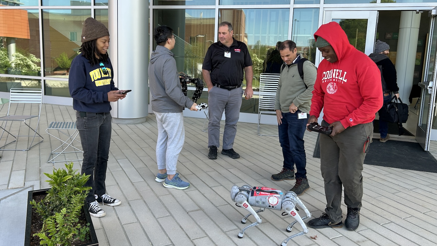 Five individuals are gathered around a small, light-colored robot dog on a paved outdoor area, with a modern building featuring large windows in the background. From left to right: a person in a dark beanie and dark sweatshirt is looking at a phone; a person in a grey hoodie and light grey pants is holding a device and looking at the robot; a man in a black polo shirt and grey pants is standing and looking at the robot; another man in a green long-sleeved shirt and jeans is looking down at the robot; and a person in a red "CORNELL UNIVERSITY" hoodie and dark pants is also looking down at the robot, possibly interacting with a device. The robot itself is four-legged and appears to be in a crouching position. There are some chairs and planters visible on the left.
