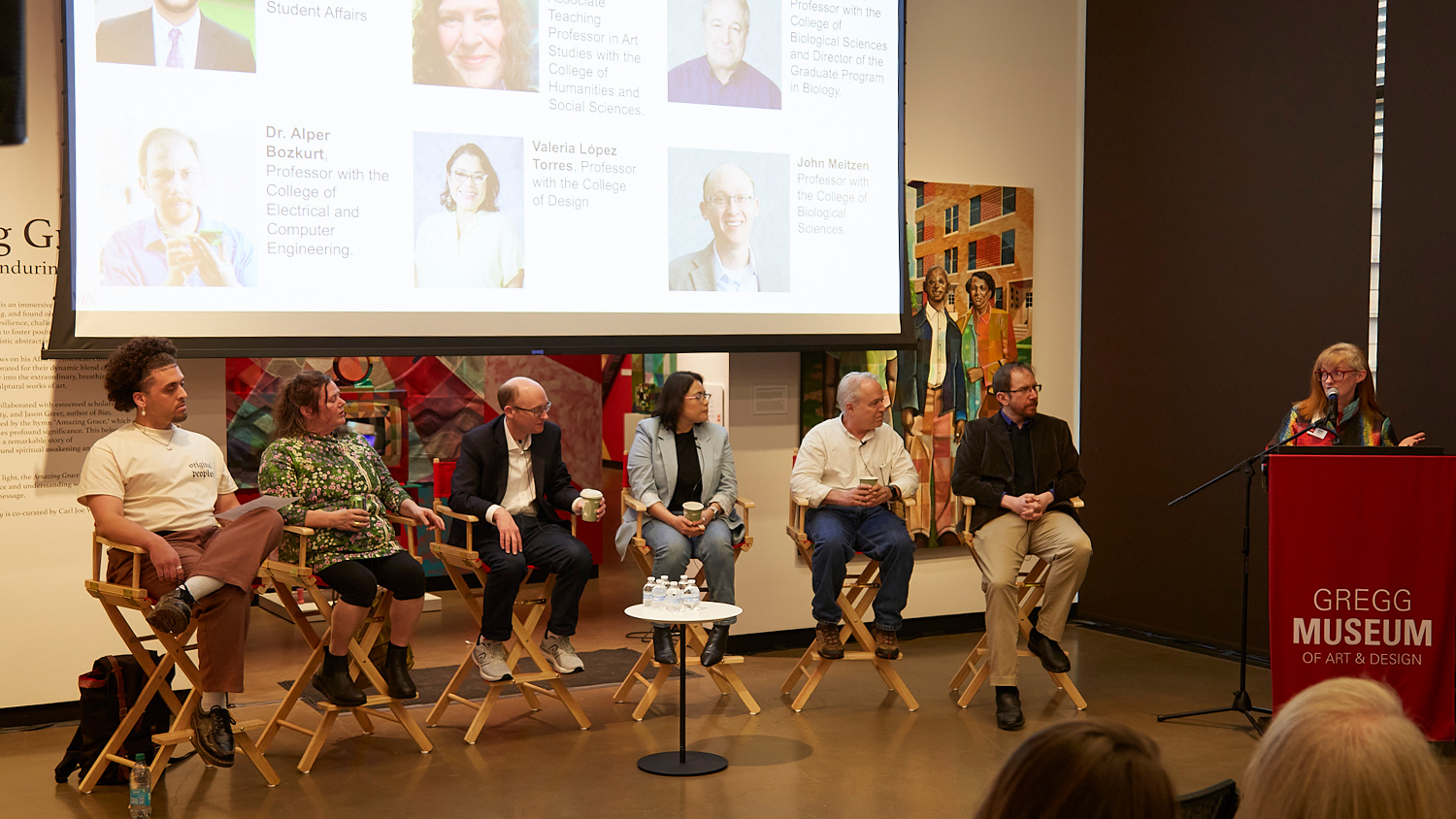 A panel of six people, five men and one woman, are seated on tall wooden stools facing an audience, with a woman standing at a red podium on the far right. Behind them, a large screen displays headshots and names of individuals, including "Dr. Alper Bozkurt" and "Valerie Lopez Torres." The setting appears to be a museum or gallery, with artwork visible on the walls and a "GREGG MUSEUM OF ART & DESIGN" logo on the podium.