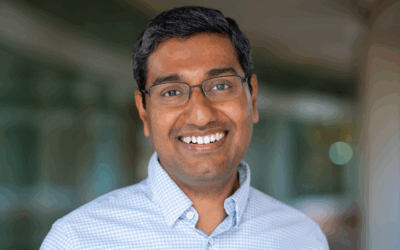 A close up photo of a smiling, brown-skinned man with glasses and dark hair. He is wearing a blue button down and standing in front of a blurry background outside.