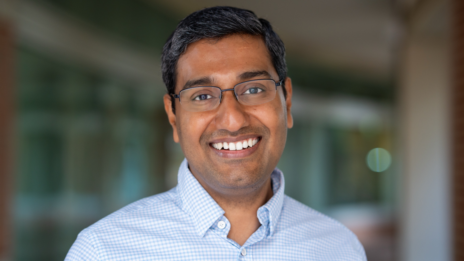 A close up photo of a smiling, brown-skinned man with glasses and dark hair. He is wearing a blue button down and standing in front of a blurry background outside.