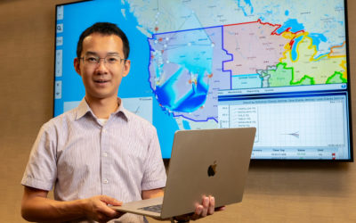 A professional headshot of a smiling man, likely an engineer or researcher, wearing a light-colored button-up shirt and glasses, holding an open MacBook laptop. He stands in front of a large monitor displaying a map of the United States and Southern Canada with lines overlaying the electrical grid and various areas shaded blue, representing real-time frequency or power data. A panel on the right side of the screen shows a data table with values like "Freq" and "Unit," indicating a system for monitoring and reporting power grid conditions.