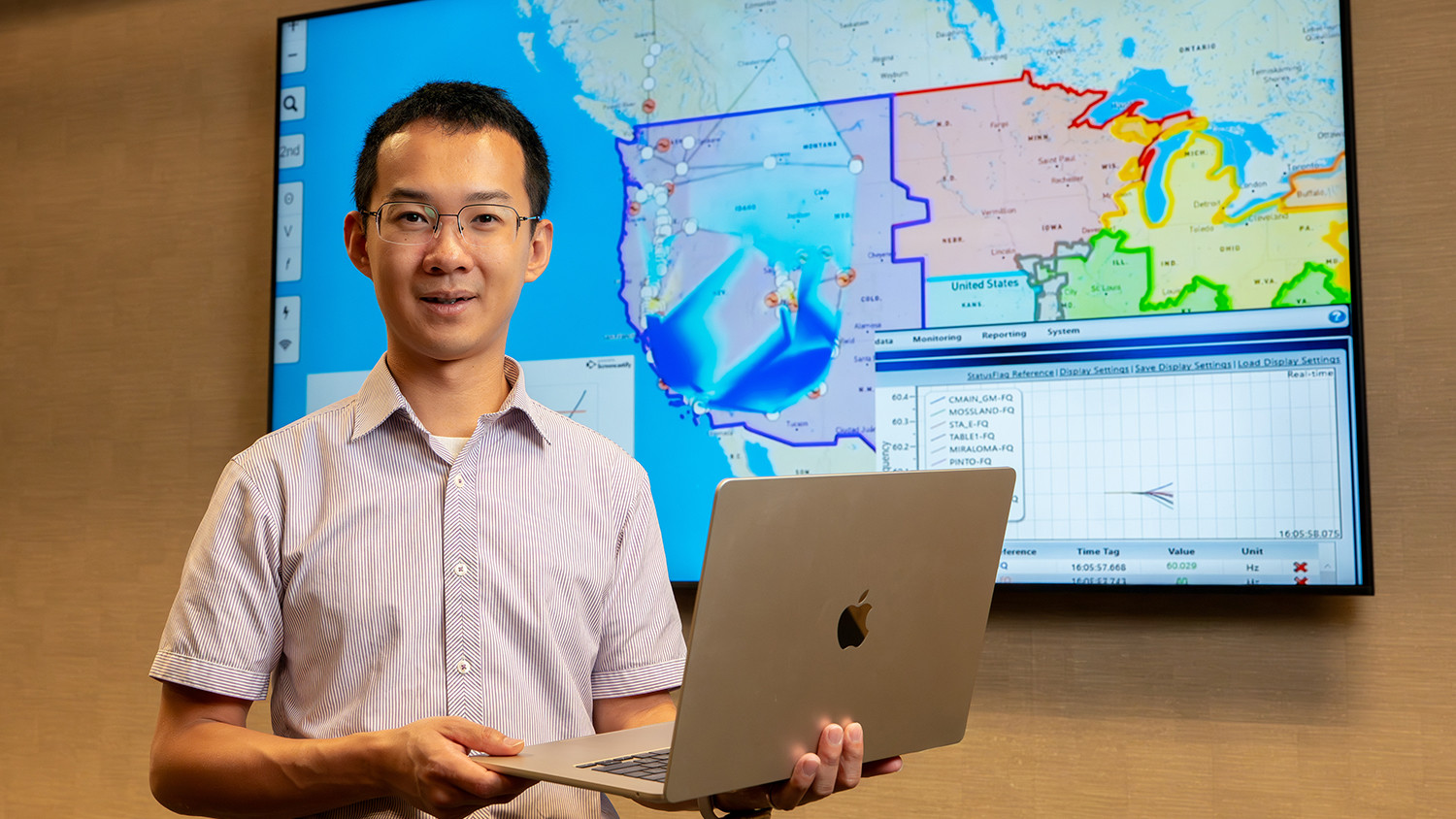 A professional headshot of a smiling man, likely an engineer or researcher, wearing a light-colored button-up shirt and glasses, holding an open MacBook laptop. He stands in front of a large monitor displaying a map of the United States and Southern Canada with lines overlaying the electrical grid and various areas shaded blue, representing real-time frequency or power data. A panel on the right side of the screen shows a data table with values like