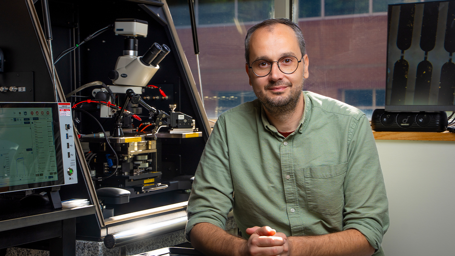 A professional portrait of a man, possibly a researcher or professor, wearing glasses and a light green button-down shirt, sitting in a lab. Behind him is complex scientific equipment, including a microscope and monitors showing data.