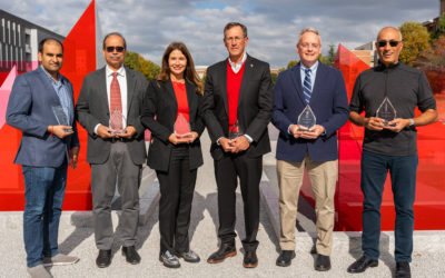 Six people, four men and two women, dressed in business casual attire, stand outdoors holding glass awards. They are positioned in front of a large, modern red and gray architectural display.
