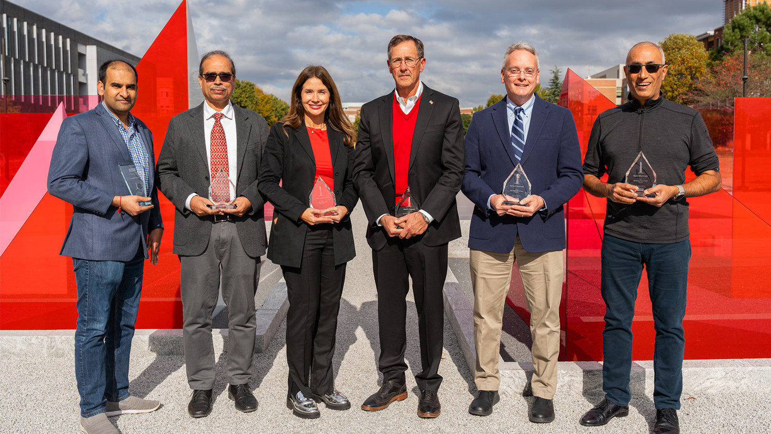 Six people, four men and two women, dressed in business casual attire, stand outdoors holding glass awards. They are positioned in front of a large, modern red and gray architectural display.