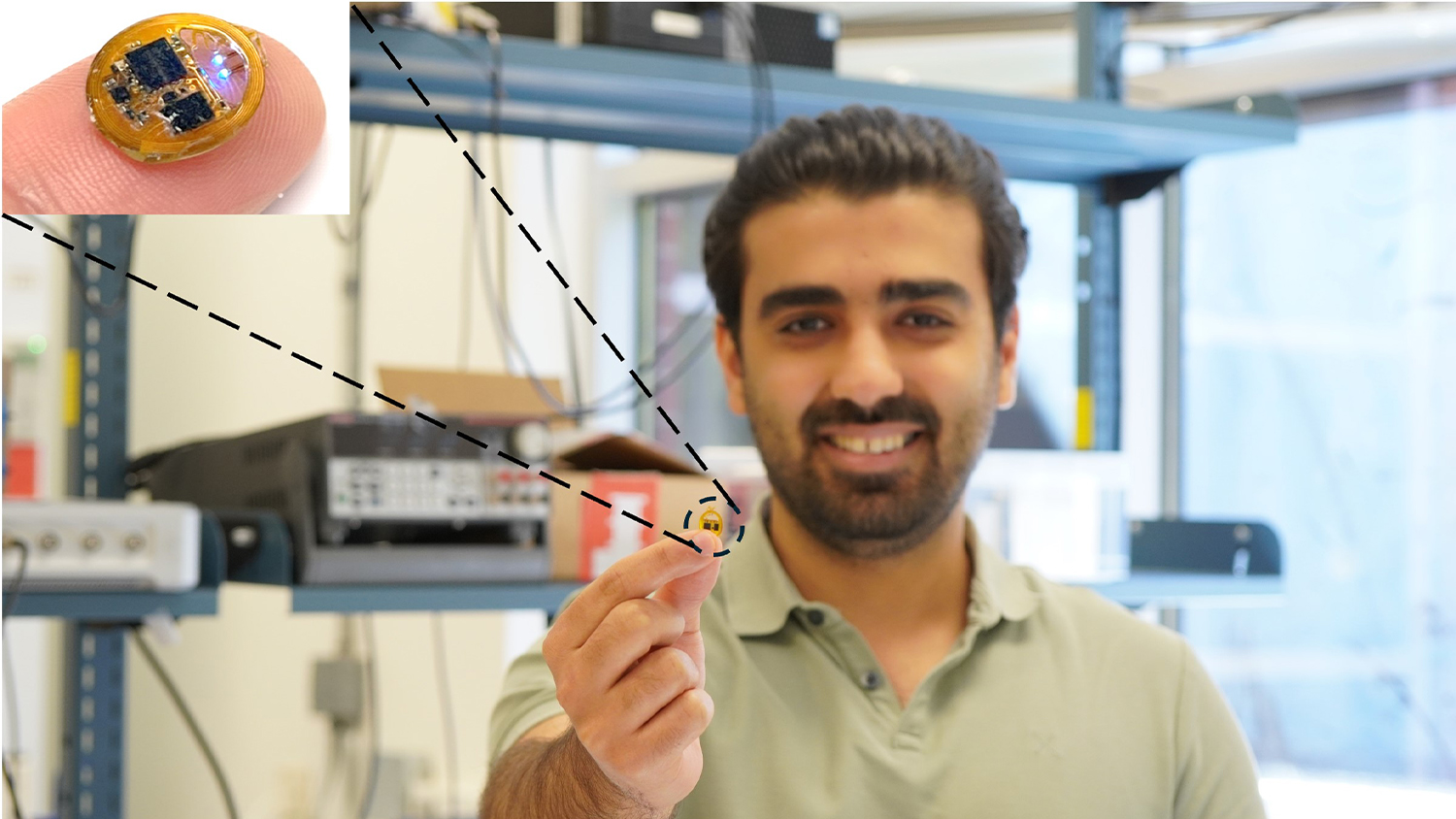 This image features a smiling man in a light green polo shirt, standing in what appears to be a laboratory or workshop setting with equipment visible on shelves behind him. He is holding a tiny, gold-colored electronic device between his thumb and index finger, extending it towards the camera.