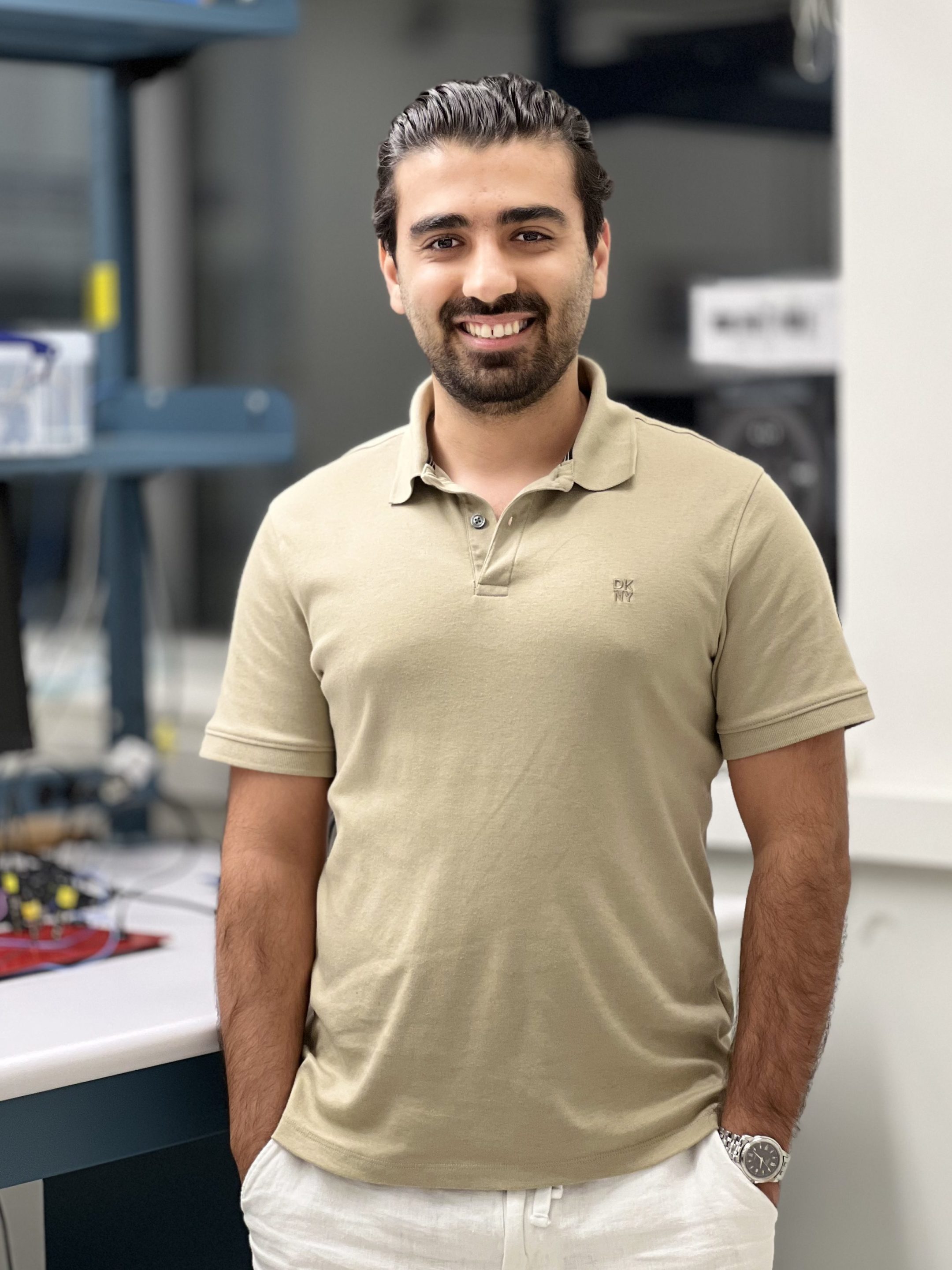 A young man with dark hair and a beard is standing and smiling directly at the camera. He is wearing a khaki/tan polo shirt and white trousers, with his hands casually in his pockets. A watch is visible on his left wrist.

He is positioned in an indoor setting that appears to be a laboratory or workspace. A counter/desk is to his left, featuring some electronic components (including a visible red circuit board) and a computer monitor. Behind him, there are laboratory fixtures, shelves, and a window or light panel. The lighting is bright.