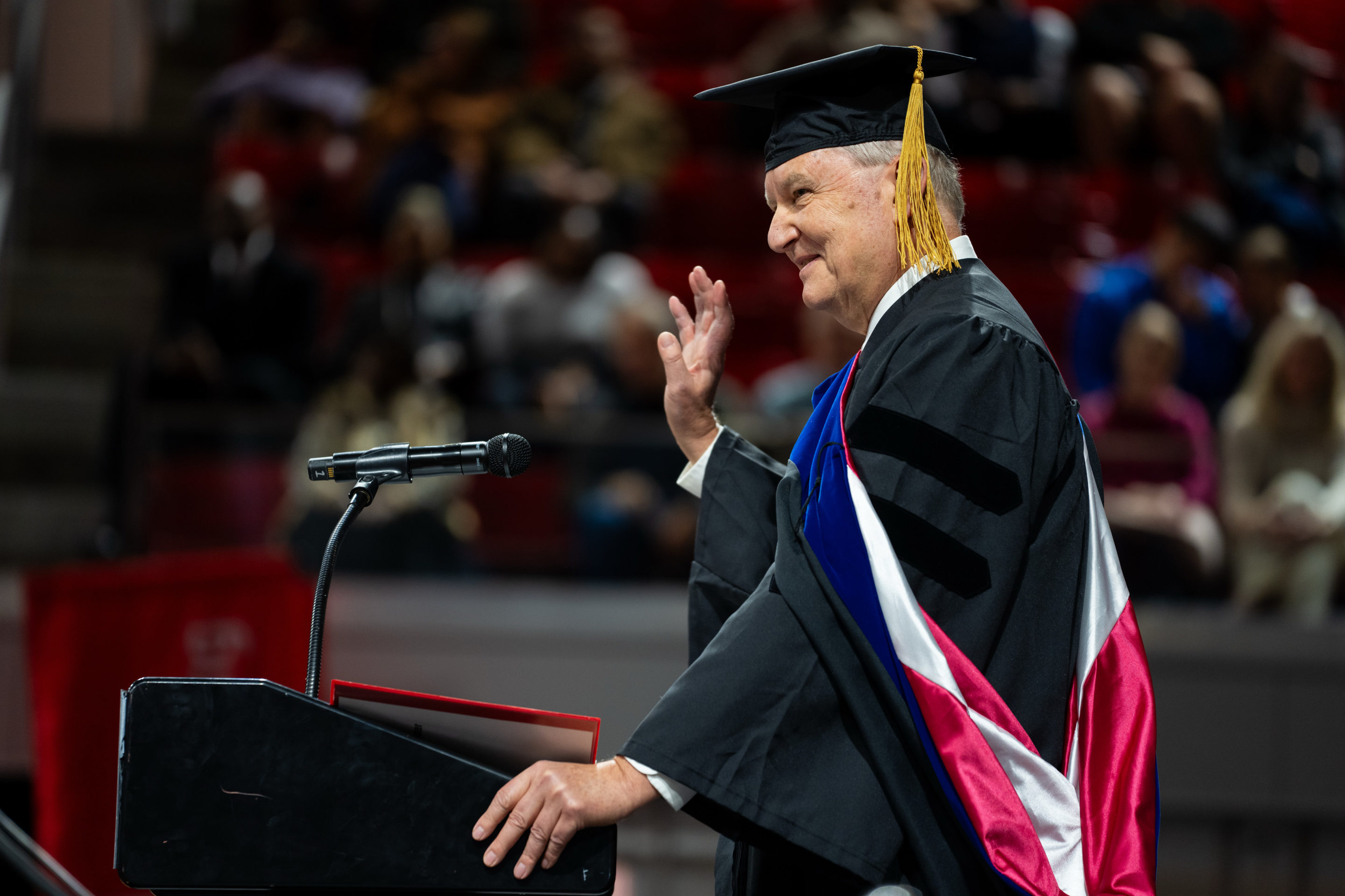 A photo of an older man, speaking at a lectern during graduation. He is wearing dark robes and a cap.