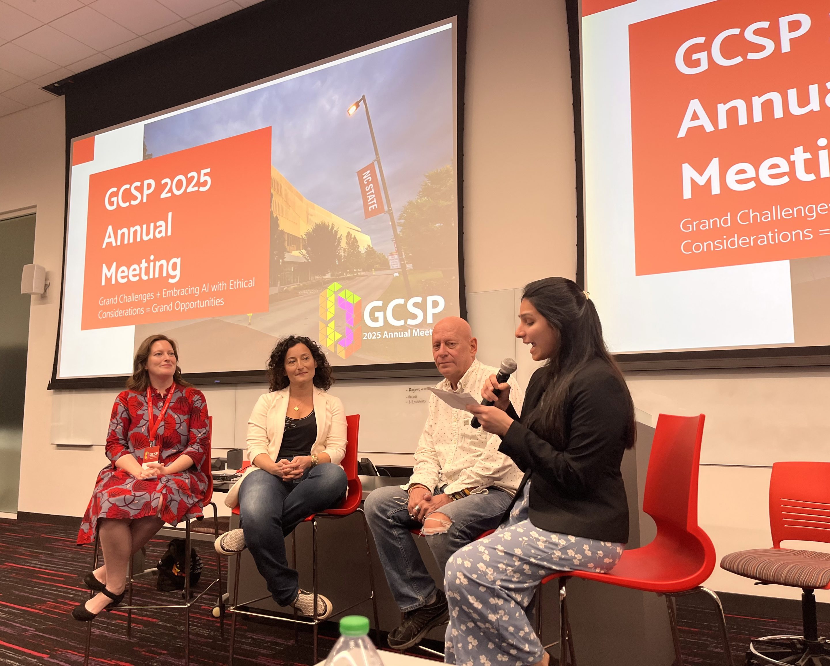 A panel discussion taking place in a conference room. Four people sit in red modern chairs on a stage in front of a large projection screen displaying "GCSP 2025 Annual Meeting". Ishita Pai Raikar, seated on the far right, speaks into a handheld microphone while looking at notes. The other three panelists, two women and one man, listen attentively.