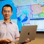 Individual holding laptop in front of a digital map display on screen, representing data analysis or geographic information.