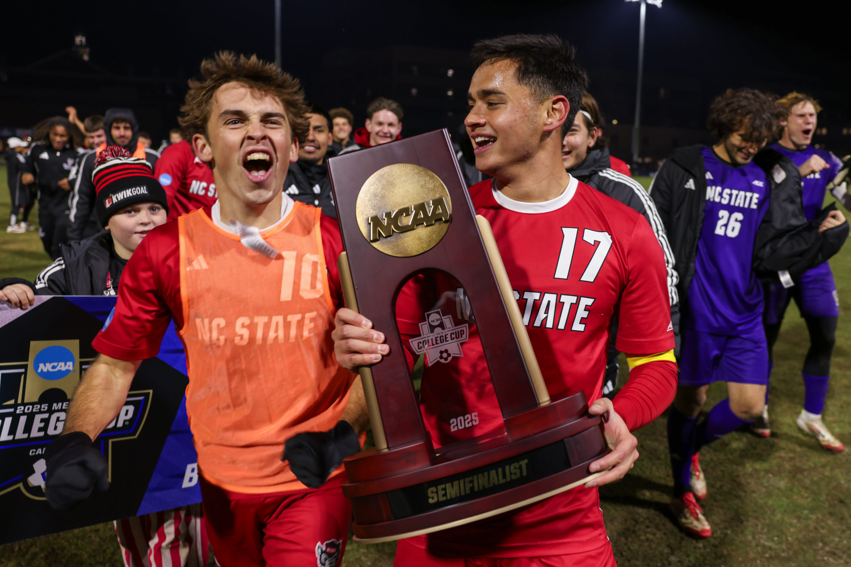 NC State soccer team celebrates with NCAA semifinalist trophy on the field at night.