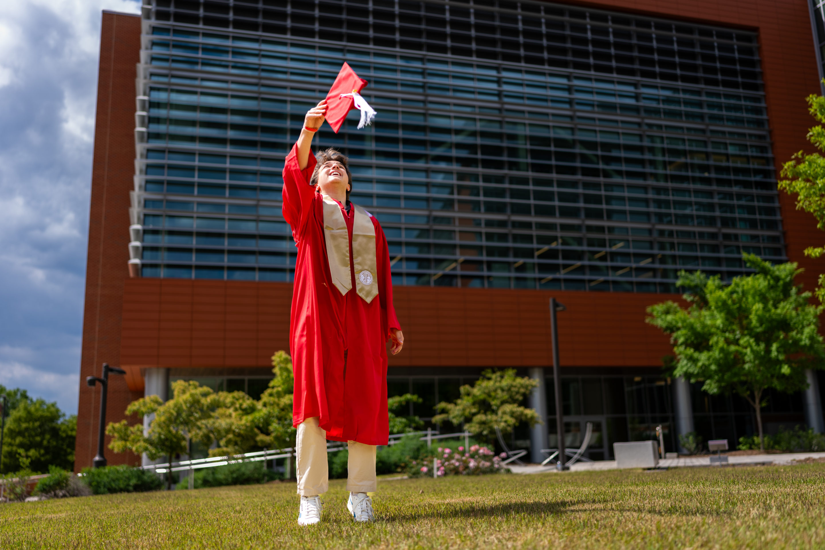 Graduate in red gown celebrates outside a modern building, raising cap in achievement against a cloudy sky.