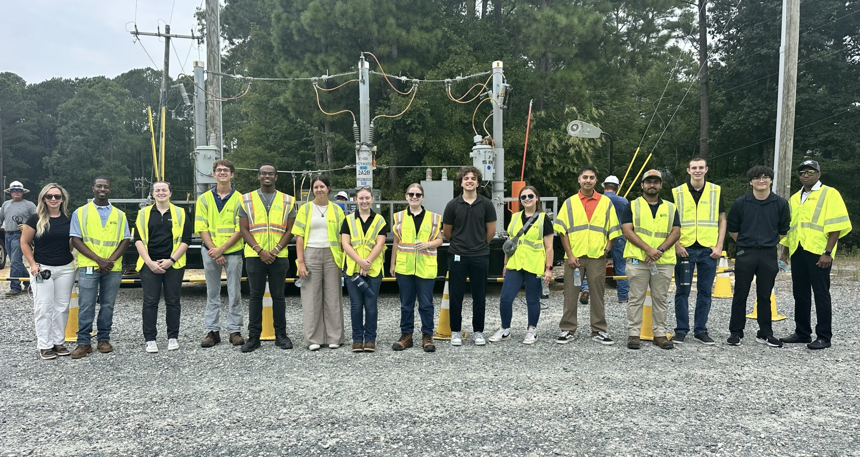 Group of utility workers and engineers in safety vests, standing outdoors near electrical equipment and trees.