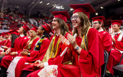 Graduates in red gowns celebrating during a commencement ceremony in a packed auditorium.