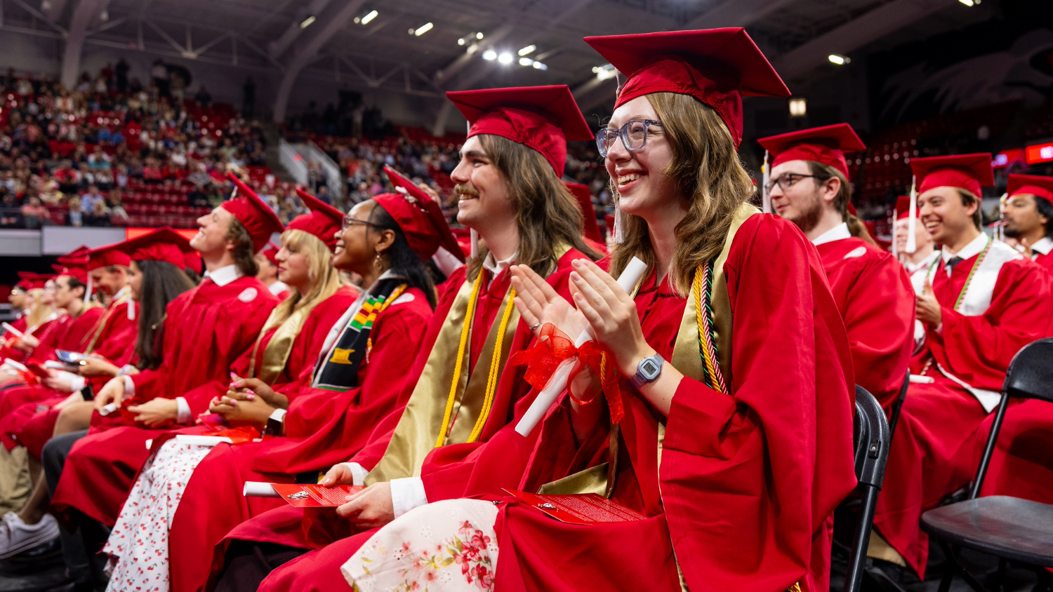 Graduates in red gowns celebrating during a commencement ceremony in a packed auditorium.