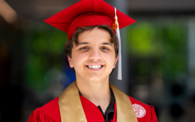 Smiling graduate in red cap and gown with gold stole, celebrating academic achievement.