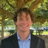 Smiling person in a suit under a tree, outdoors, on a sunny day.