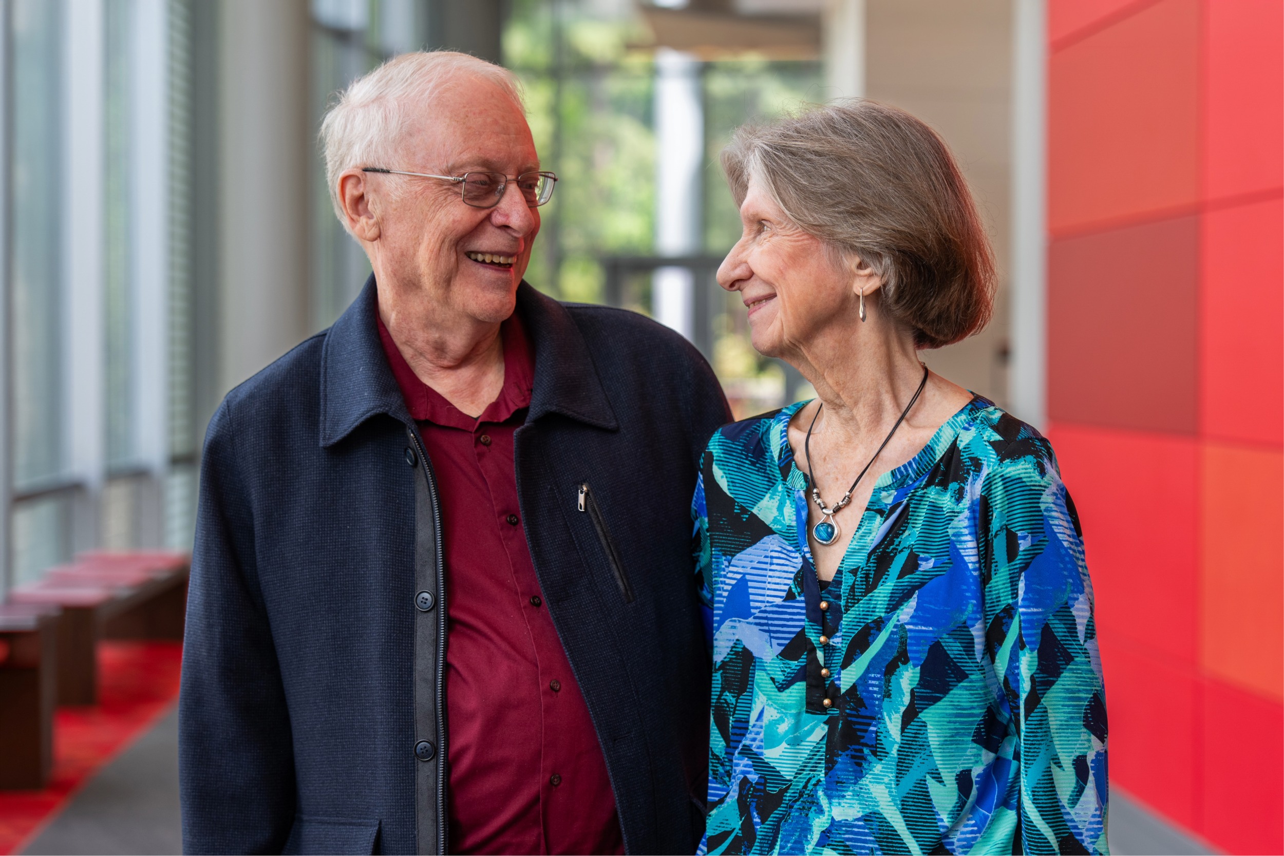 Elderly couple smiling at each other in a bright hallway with red and blue accents.