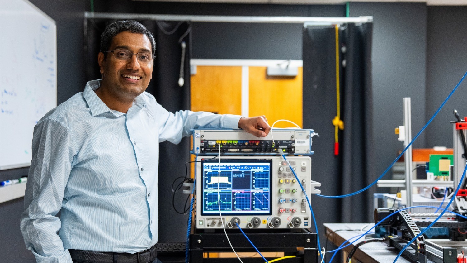 Scientist in lab with advanced equipment, smiling and standing beside an electronic testing device.