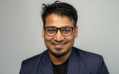 Person smiling, wearing glasses and a blazer against a neutral background.