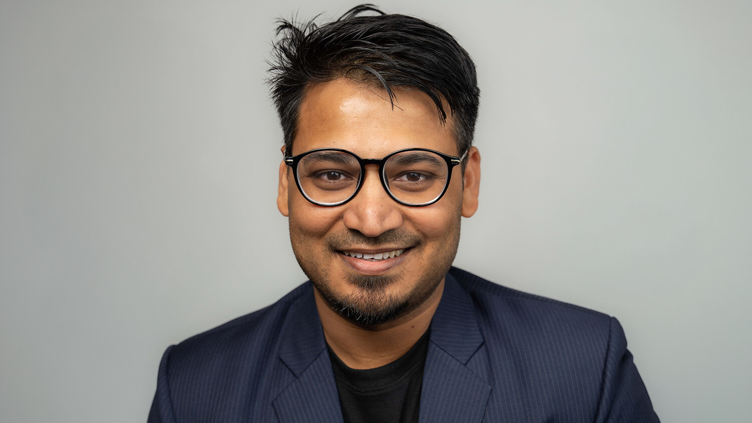 Person smiling, wearing glasses and a blazer against a neutral background.