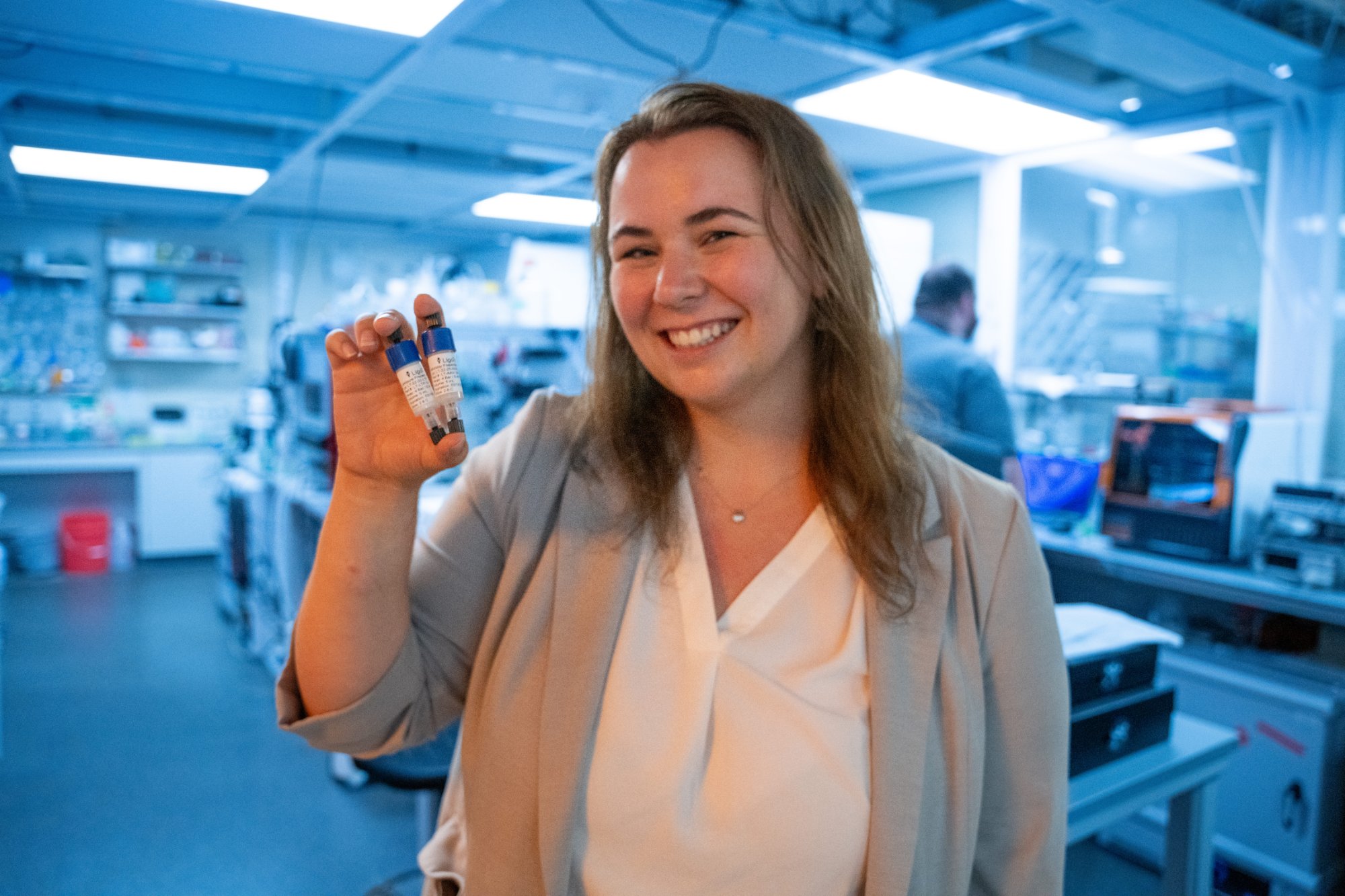 A researcher smiles and holds up two small sample vials in a lab.